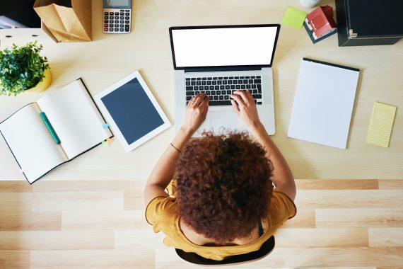 Overhead shot of a young lack woman typing on a silver laptop ins clean modern office space.