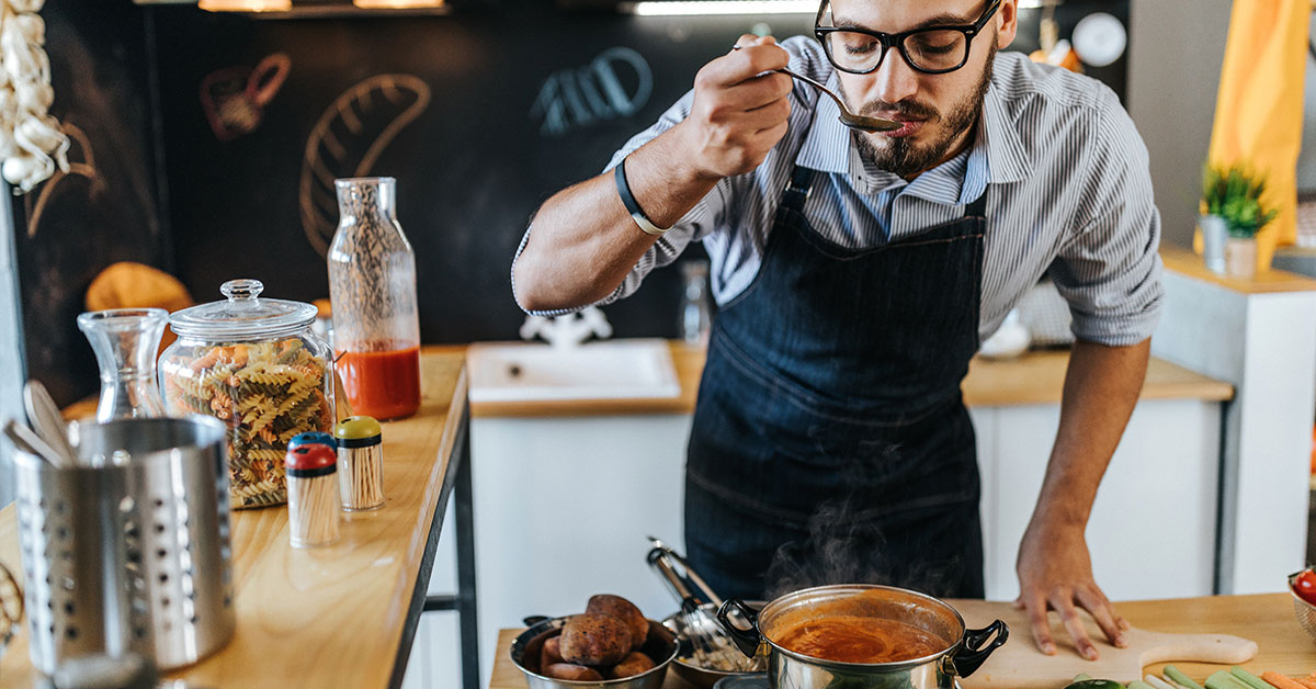 A white mid-30s man in a modern kitchen tasting soup he is preparing, surrounded by fresh ingredients.