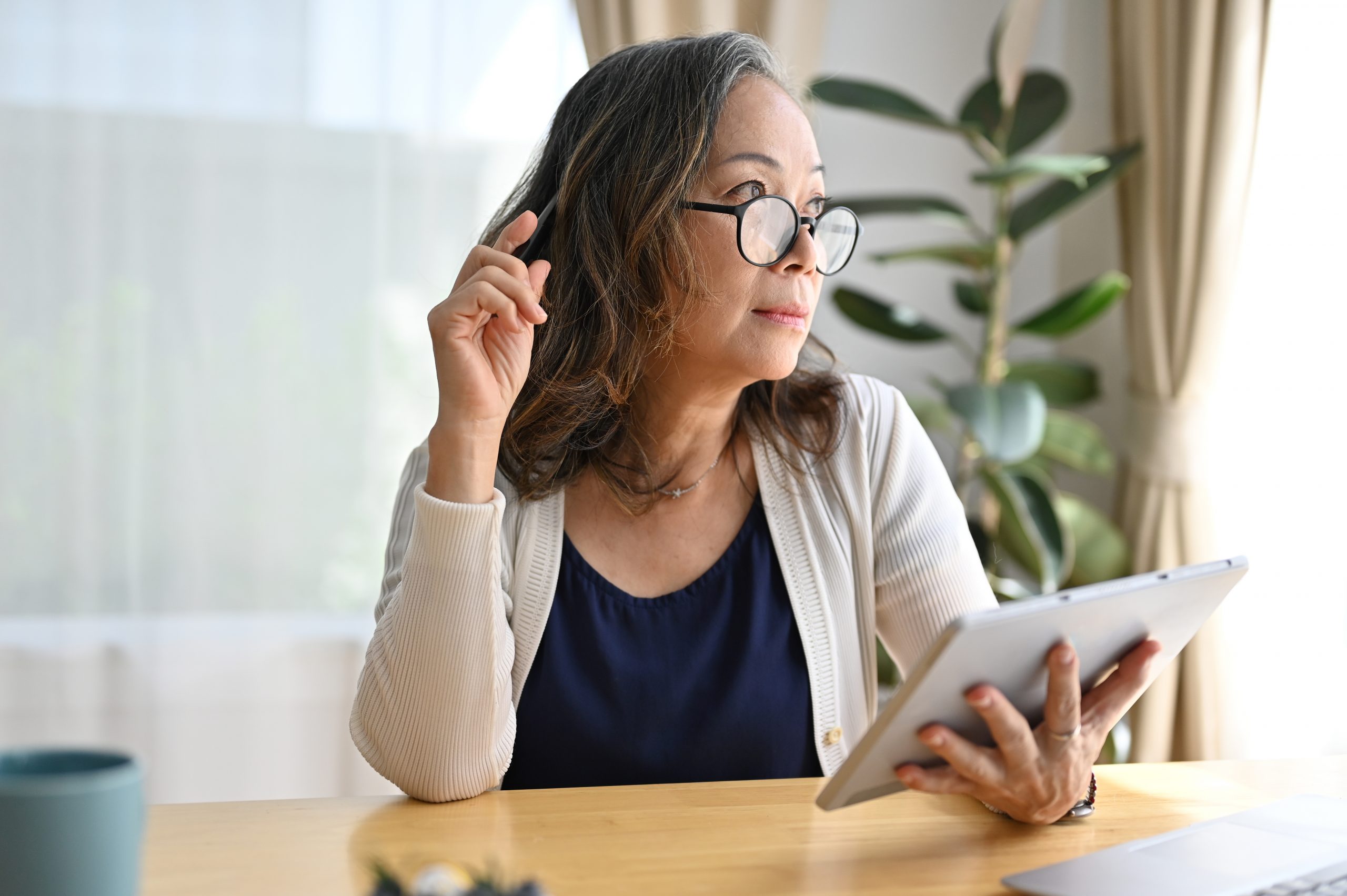 A mature south-east Asian woman using a digital tablet touchpad in a sunlite room, pensive thinking some ideas and looking through the window.