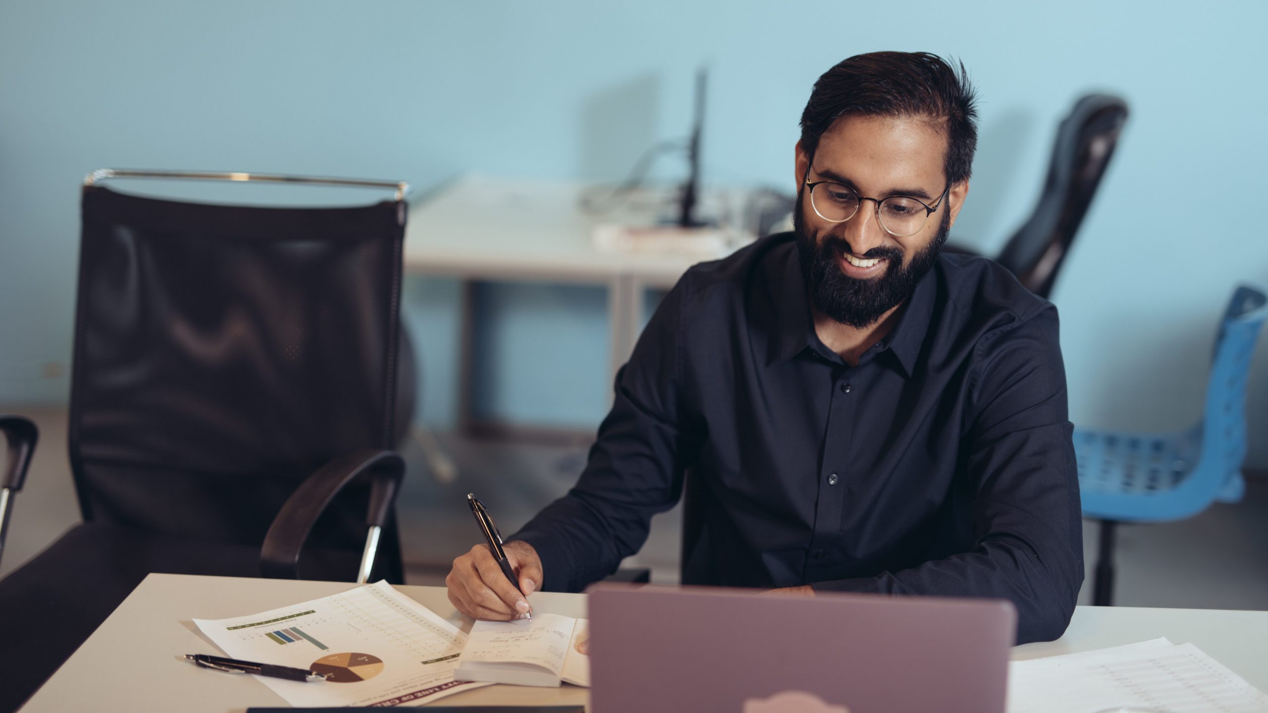 South Asian man in dark button up shirt with beard, sit smiling and at desk while notetaking and viewing a laptop screen