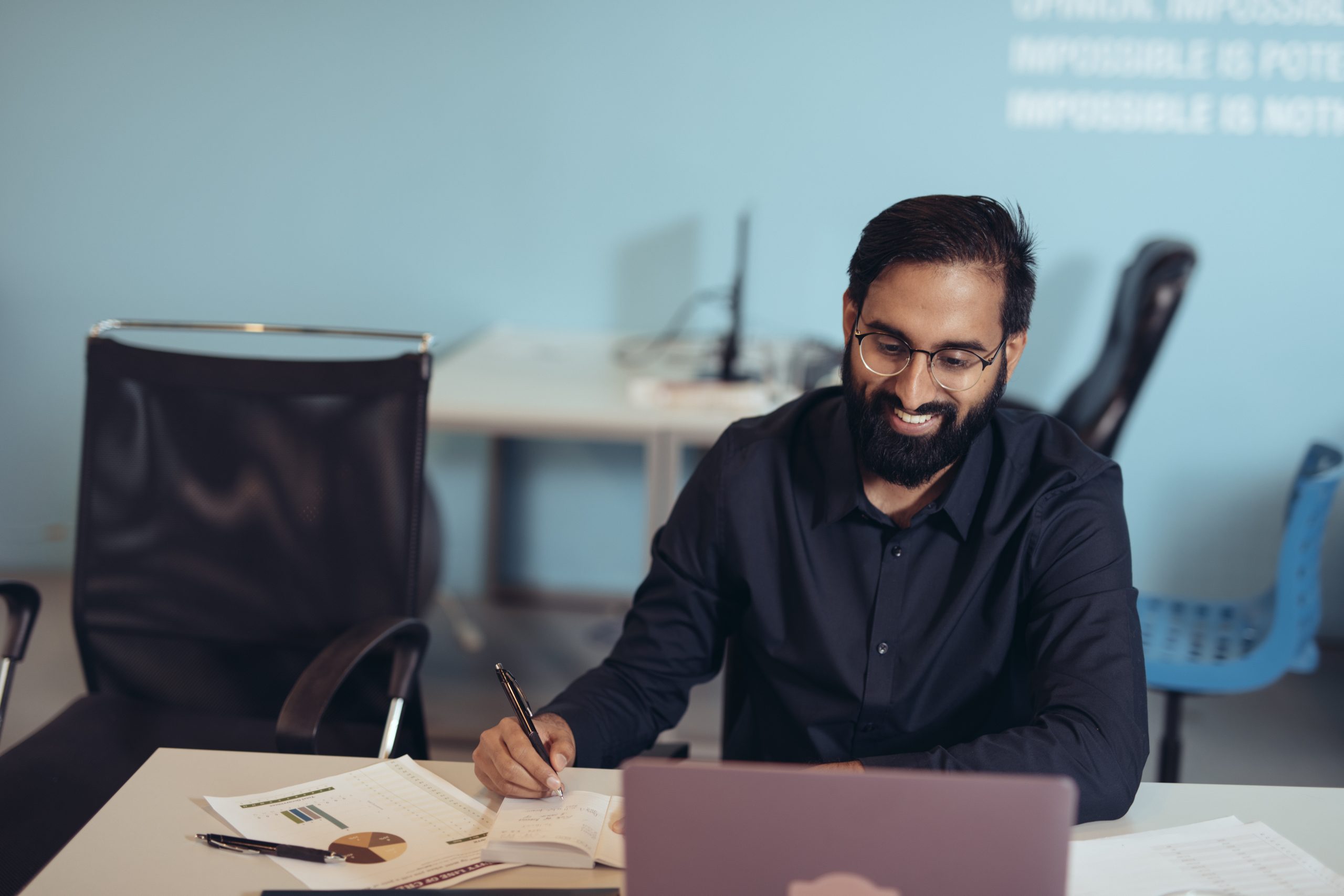 South asain man in dark button up shirt with beard, sit smiling and at desk while notetaking and viewing a laptop screen
