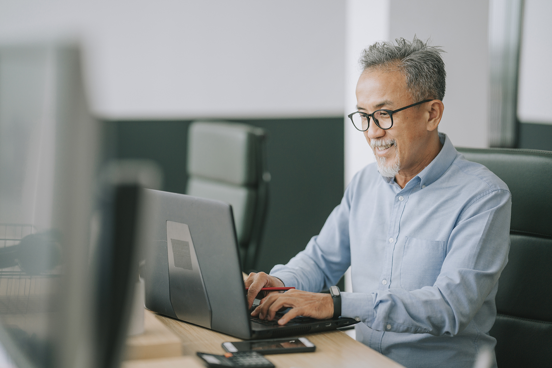 Smiling senior East Asian man typing on a laptop in a modern office space