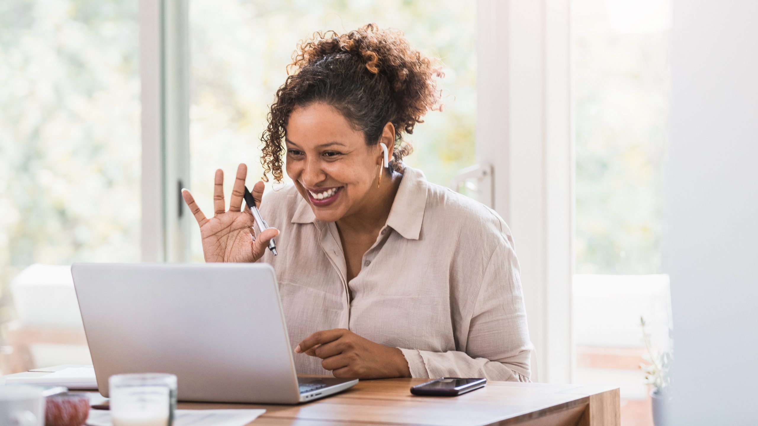 A smiling Black woman, in a bright and airy room, waves to the camera of her laptop while wearing wireless headphones