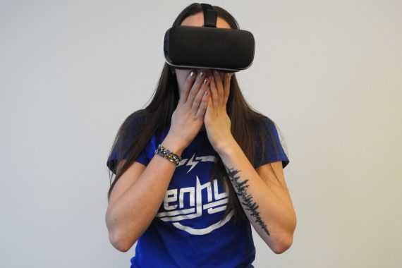 Indigenous woman with long dark brown hair wear Virtual Reality googles and blue t-shirt holds her hands up to her face in an expression of awe.