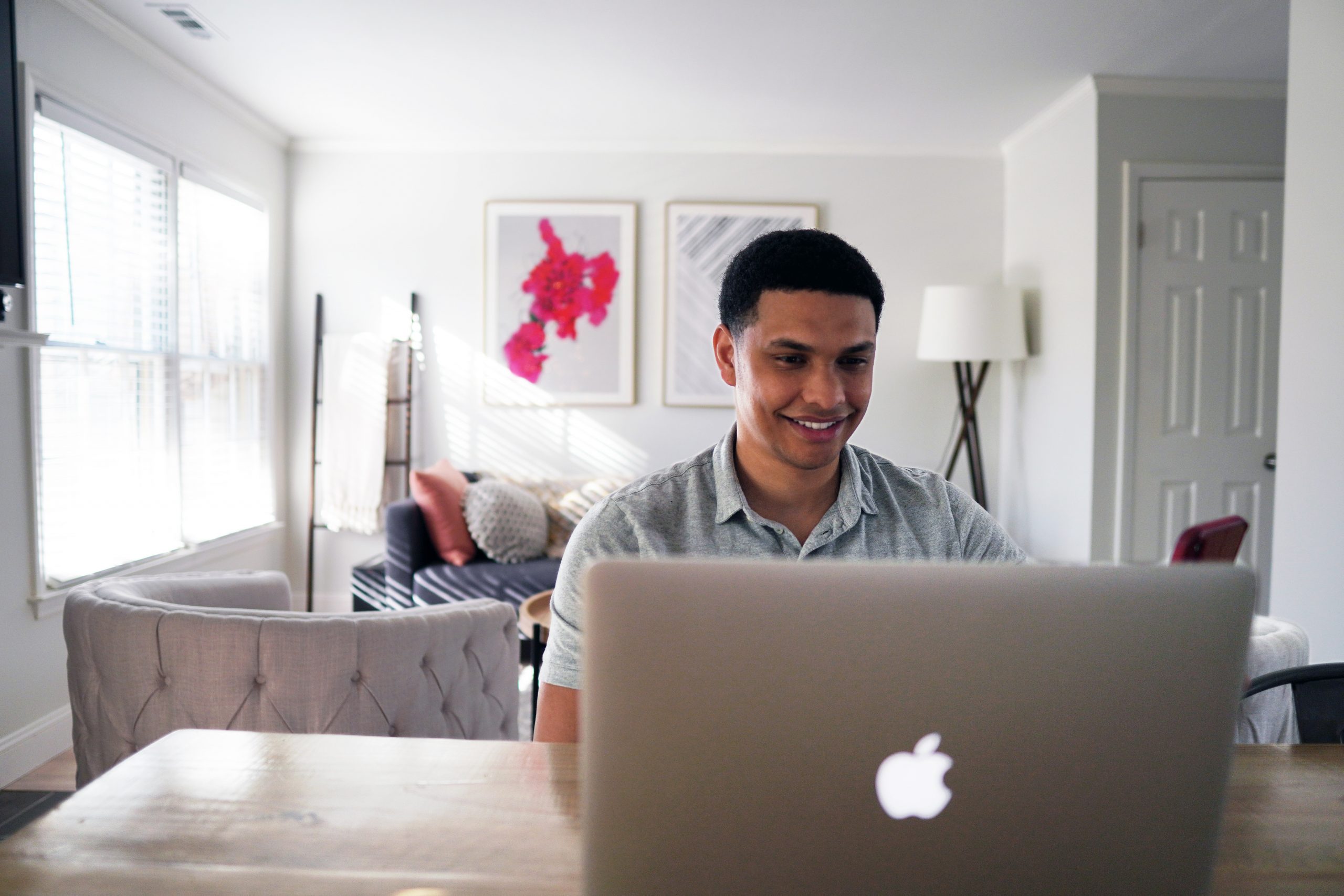 Smiling and confident mixed race  young male sits in front of laptop, behind him is a sunlight modern home
