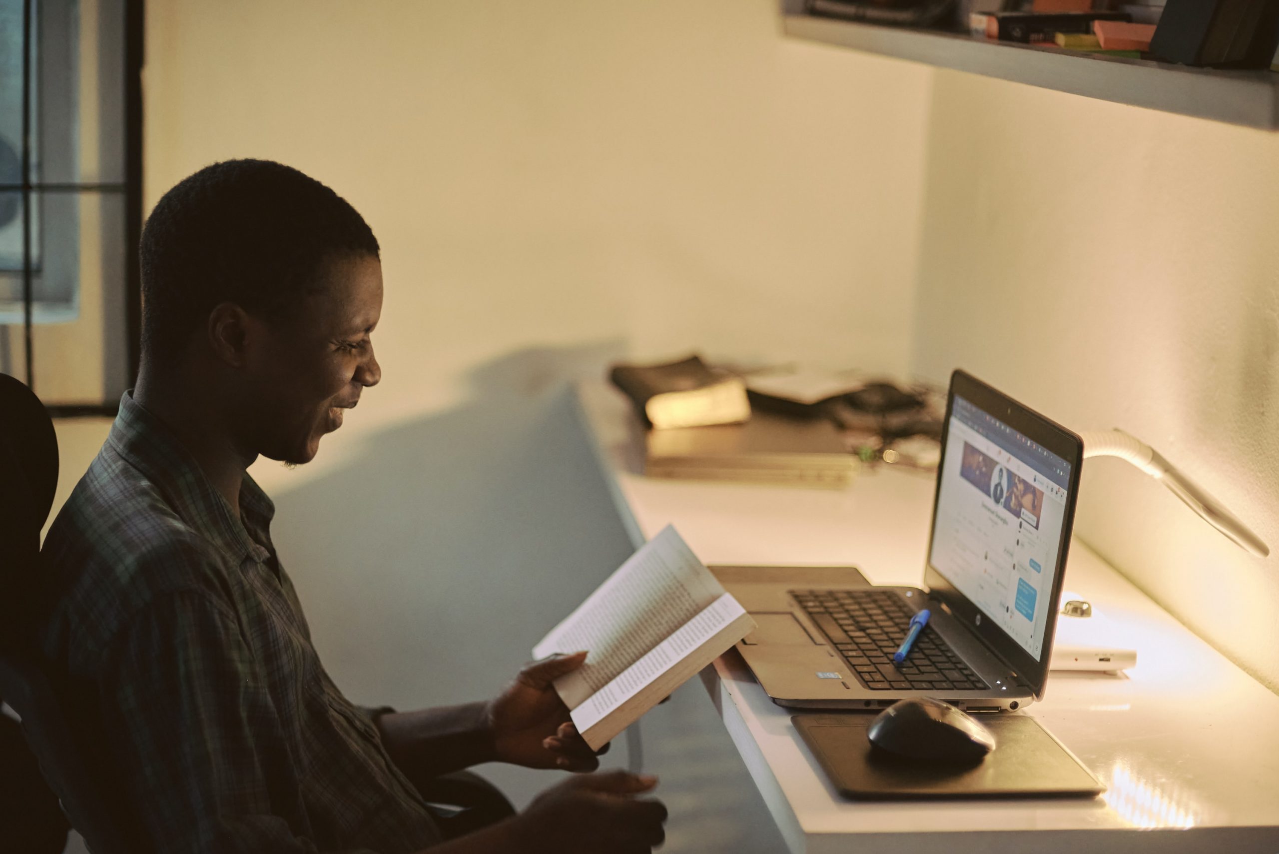 Black man contently sits in front of open laptop with open book learning.