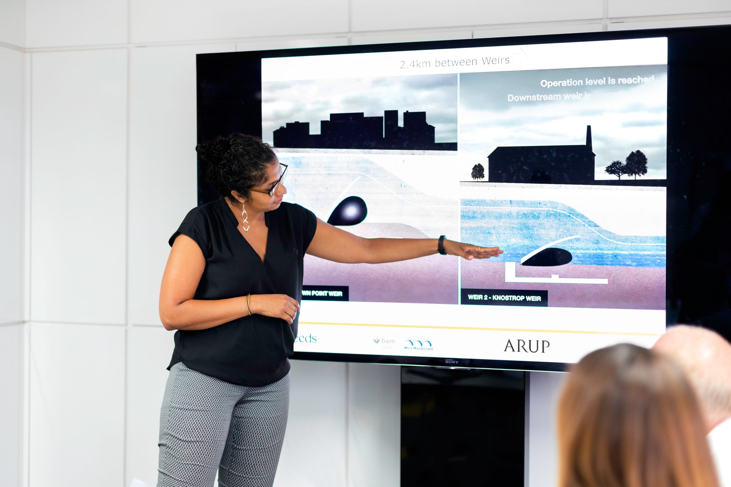 Professionally dressed South Asian woman presenting in front large digtal screen pointing the engineering diagram of a weir.