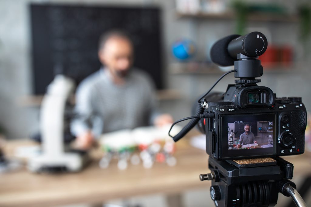 An in foucs digital camera on a tripod with attached microphone sits in front of extremely background of blurred presentor sitting and table with instructional materials sourrounding them.