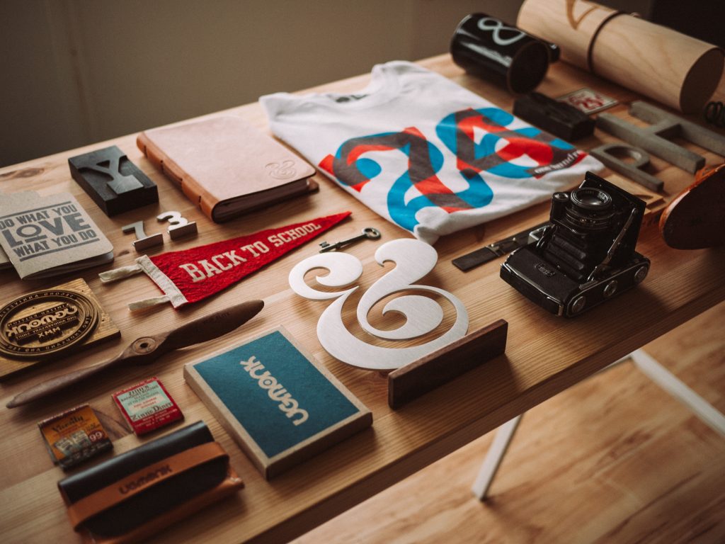 An organized set of items spread on a wooden desk, includes, anitque camera, pennate, print block, large cutout ampersand, books, notebook and propeller.
