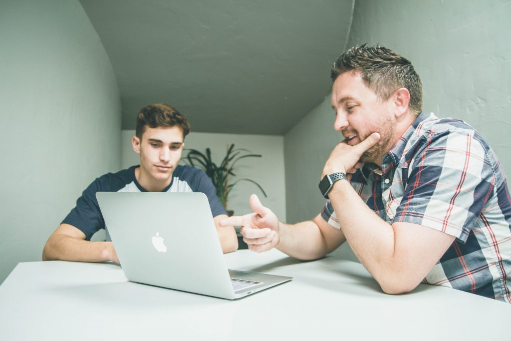 White male teenager sits and table with white male parent while review his work on laptop. the adult is pointng towards the screen.