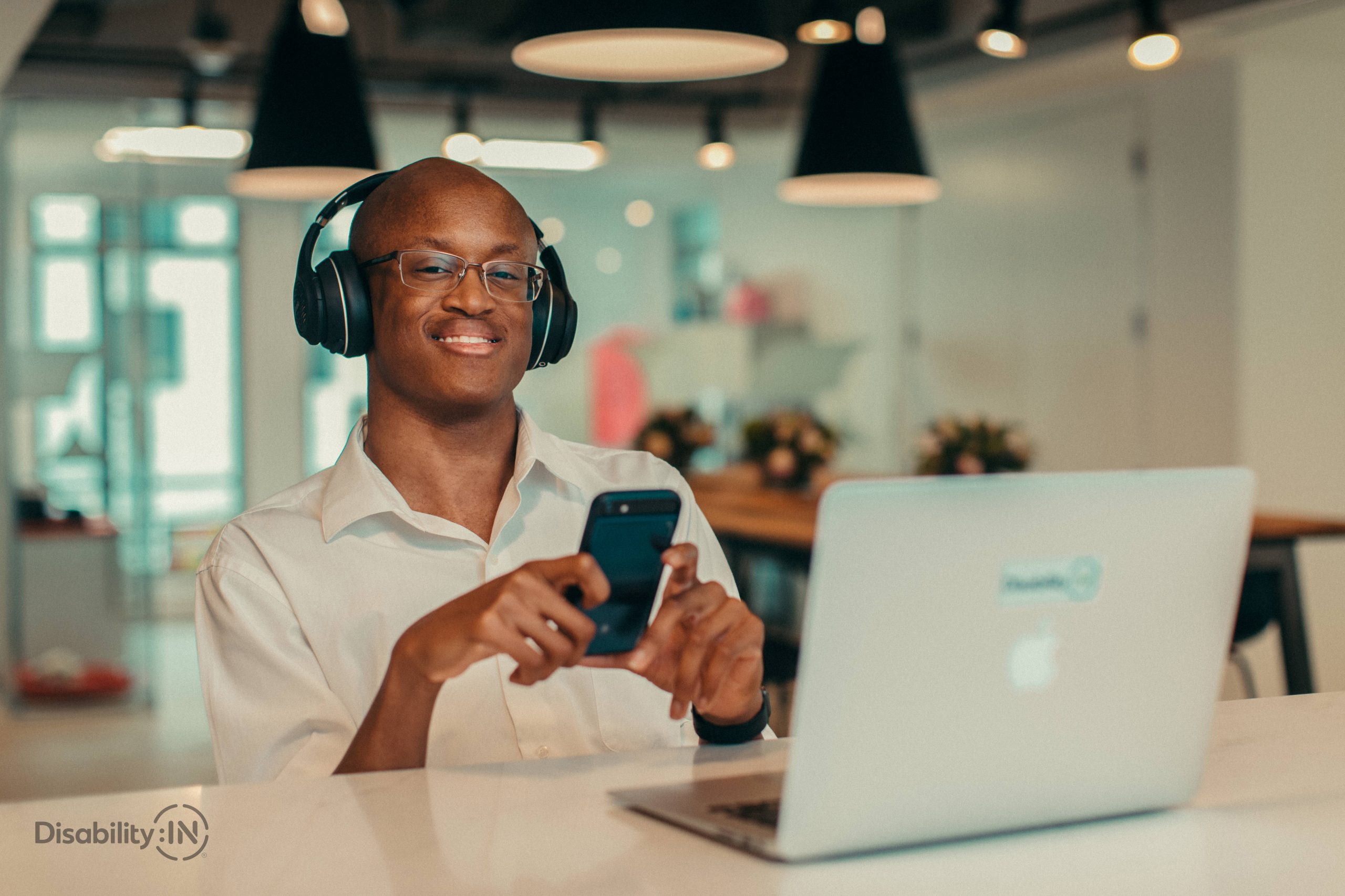 Black male wearing over the ear headphones, while listening to smartphones he is holding in his hands, sits in front of an open laptop.