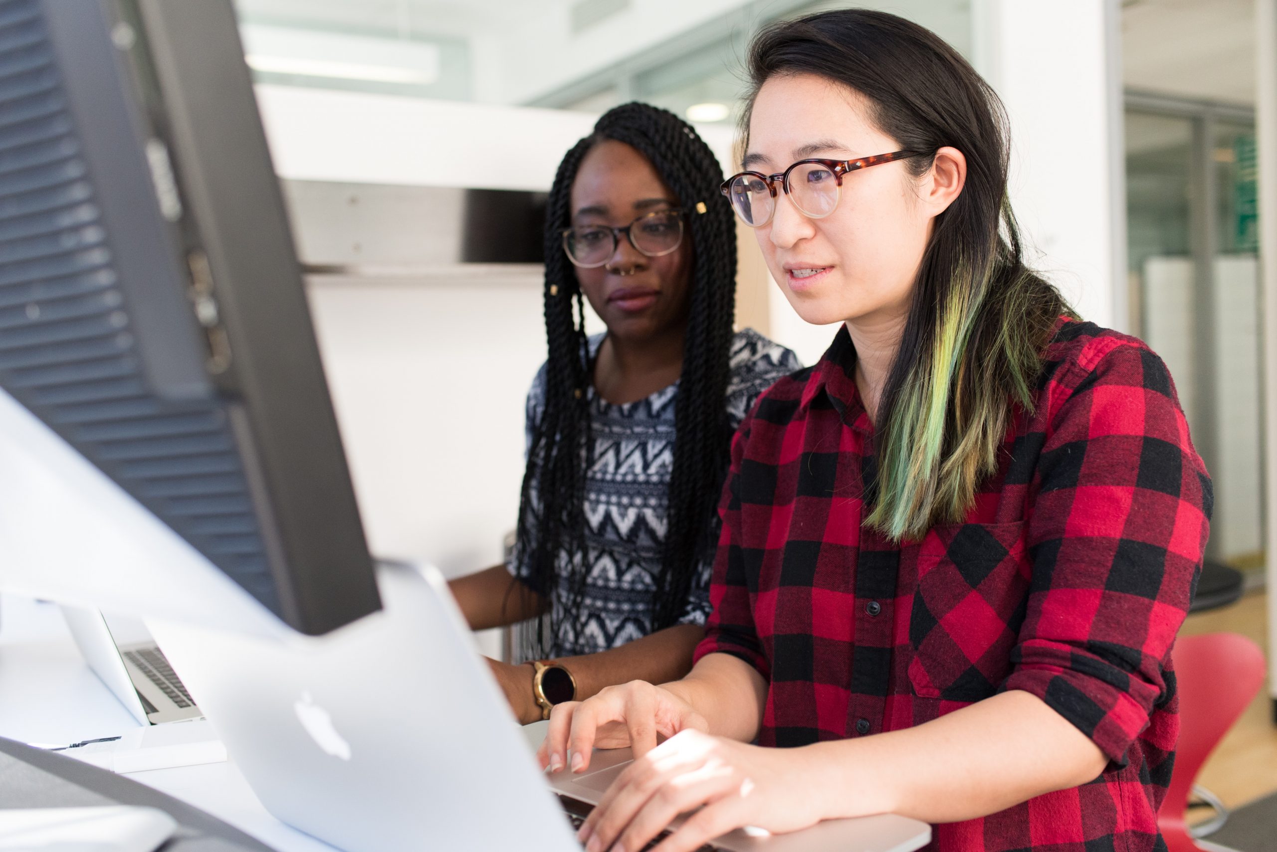 Two female presenting students one East asian and the other Black work together with laptops to solve a problem.