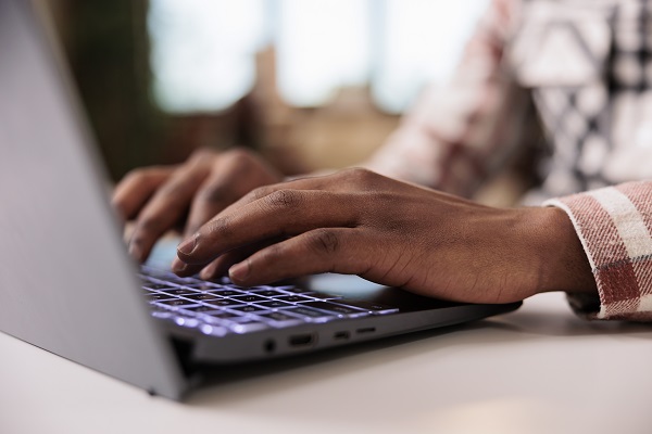close up shot of a person with darker skin tones hands type on a laptop computer keyboard.