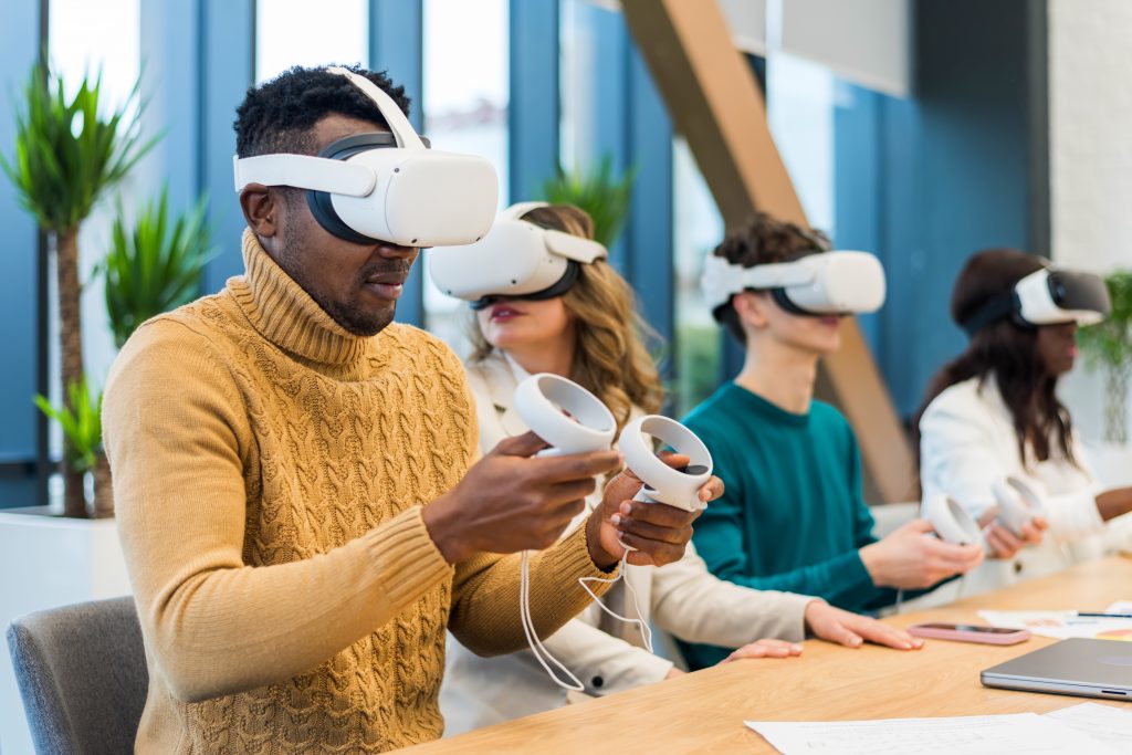 A diverse group of students all sitting along a desk wearing VR goggles and holding dual controllers.