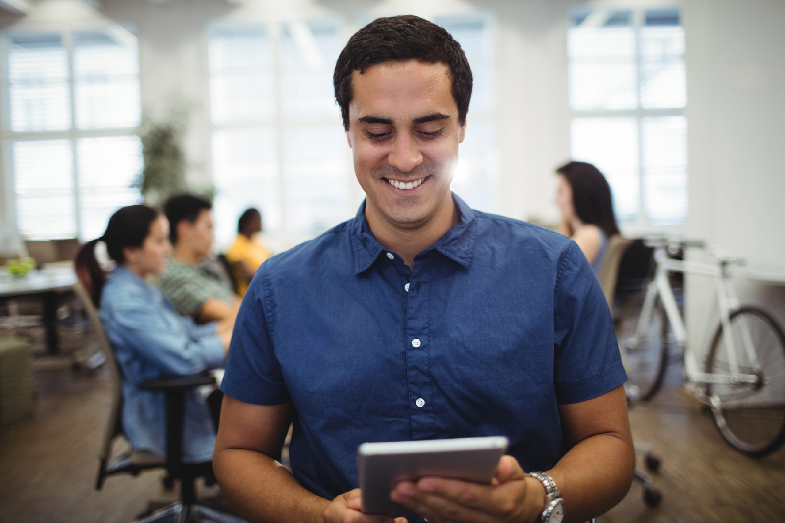 Latino male smiling while using and digital tablet, behind ho is blurred background of people sitting at a table.