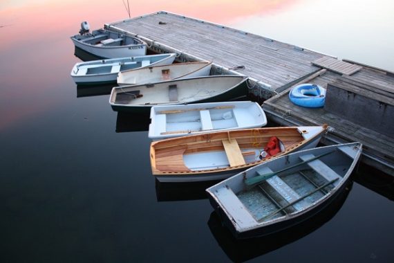 Seven two-man fishing boats, of a variety of sizes, styles, materials and ages sit tied up to a weathered wood dock on a perfectly calm lake during sunset.