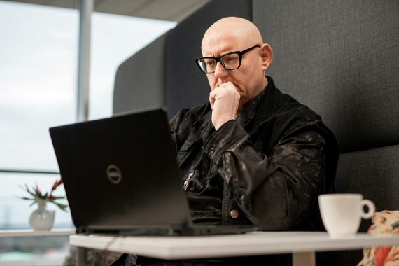 Bald white male in dark clothing staring contemplatively at his open laptop in modern cafe space.