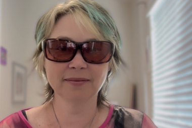 A white woman with short brown, blonde and teal streaked hair smiles and wears large dark glasses, the kind that protect eyes from light. She wears a pink top in a bright room. 